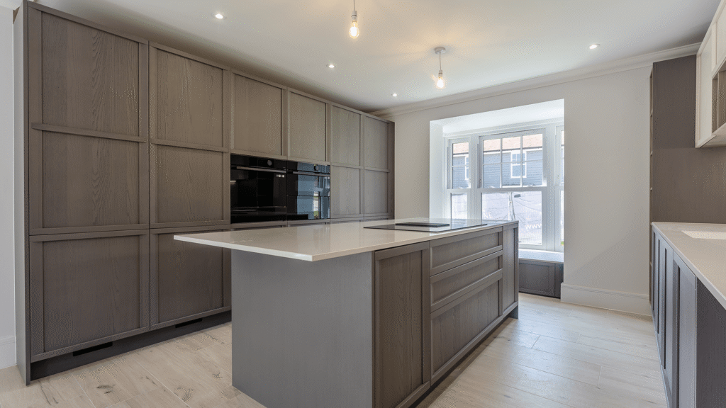 Internal image of a new builds kitchen. the floor is pale amtico, the far wall is full of storage, the kitchen is a dark grey colour. There is an island in the middle where we can see the hob and white countertops.
