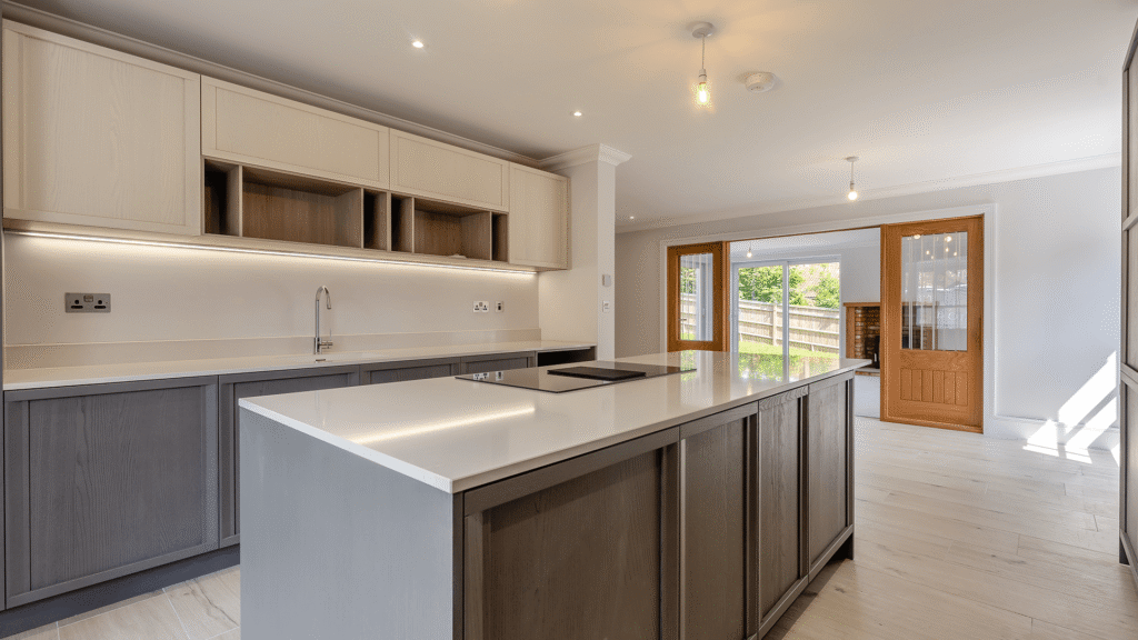 Internal image of a new builds kitchen. the floor is pale amtico, the taller cupboards are a pale wood colour and the rest of the kitchen is a dark grey colour. There is an island in the middle where we can see the hob and white countertops.