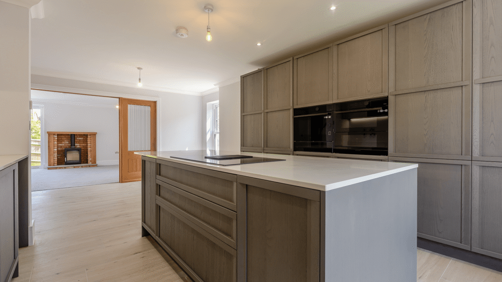 Internal image of a new builds kitchen. the floor is pale amtico, the far wall is full of storage, the kitchen is a dark grey colour. There is an island in the middle where we can see the hob and white countertops.