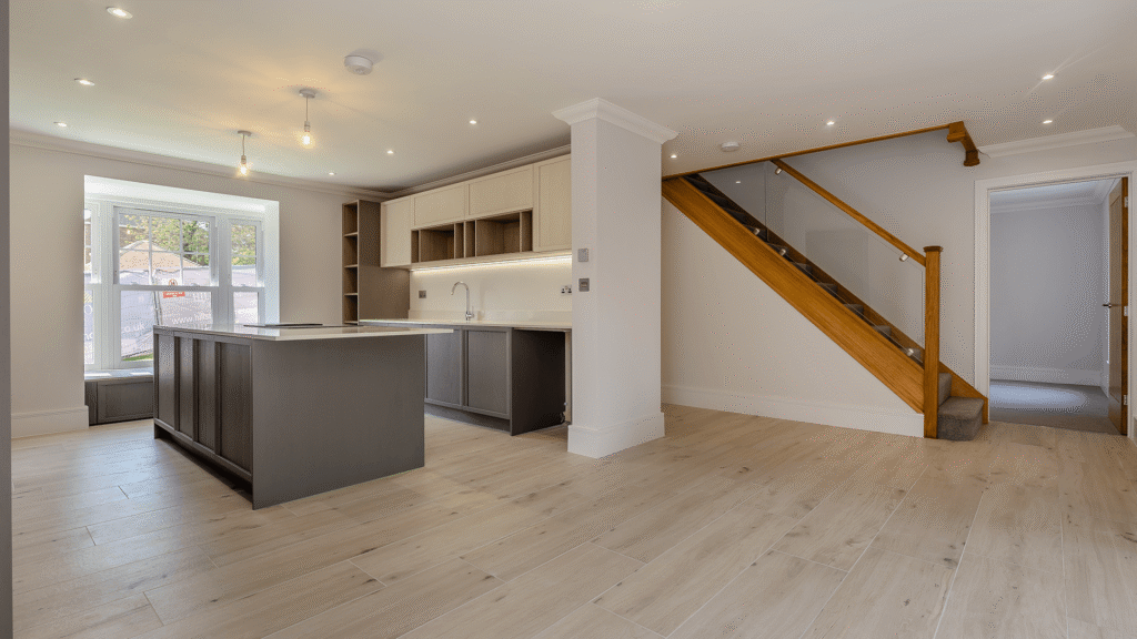 Internal image of a new builds kitchen. It is not dressed, but there is Amtico flooring and white walls, the kitchen is dark grey and the taller cupboards are a pale wood. There's a large island, and on the right we can see a dark wood staircase with dark neutral carpet on the stairs.
